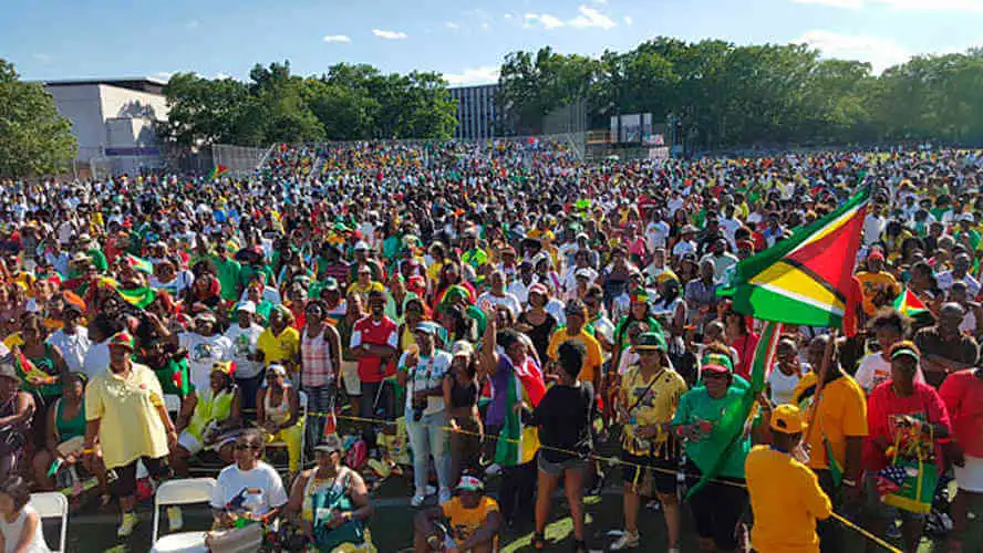 A large crowd filling a park at an outdoor Guyanese festival, the national flag flying above them